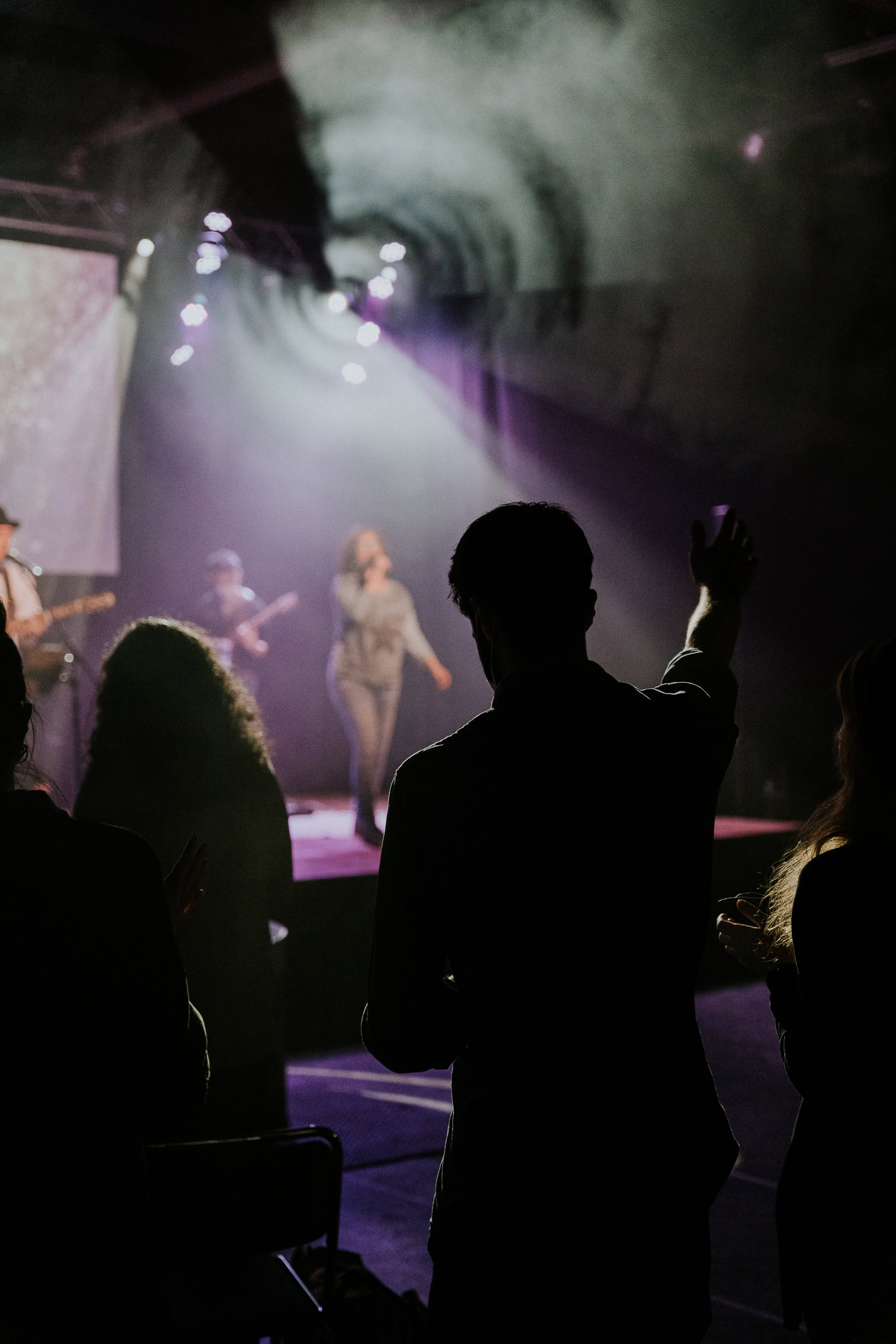 Menschen beten und feiern Gottesdienst mit Musik auf einer Bühne, im dunklen Raum mit bunten Lichtern, in der Kirche Augsburg.