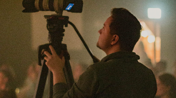 Junge Mann filmt bei einem Event mit professioneller Kamera, im Hintergrund sind unscharfe Menschen und Lichter, typische Atmosphäre eines christlichen Events in Augsburg.