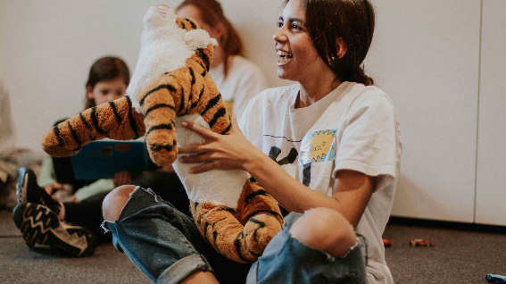 Eine Frau hält ein Tiger-Plüschtier, während sie lächelnd mit anderen Personen im Hintergrund sitzt.