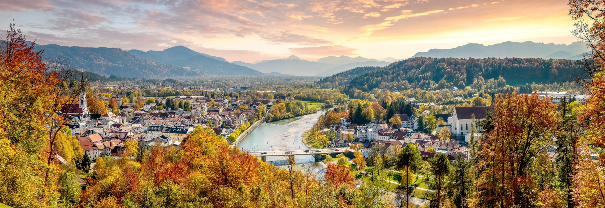 Ein Panorama der Stadt mit Fluss, umgeben von Bäumen mit Herbstfärbung, Berge im Hintergrund.