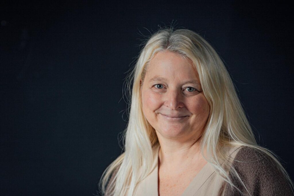 Freundliches Porträt einer Frau mit langen blonden Haaren vor dunklem Hintergrund, geeignet für christliche Gemeinde-Website.