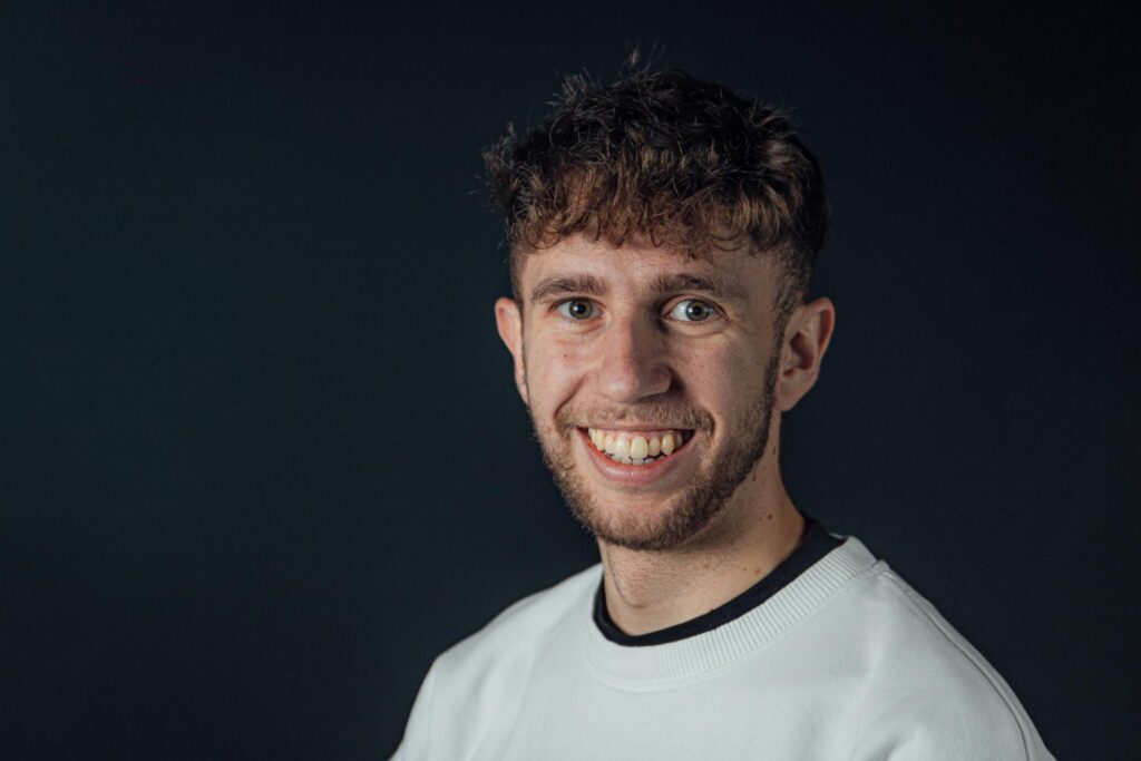 Lächelnder junger Mann mit braunen Haaren und weißem T-Shirt, professionelles Porträt vor dunklem Hintergrund, geeignet für christliche Gemeinschafts- oder Kirchenwebseiten.