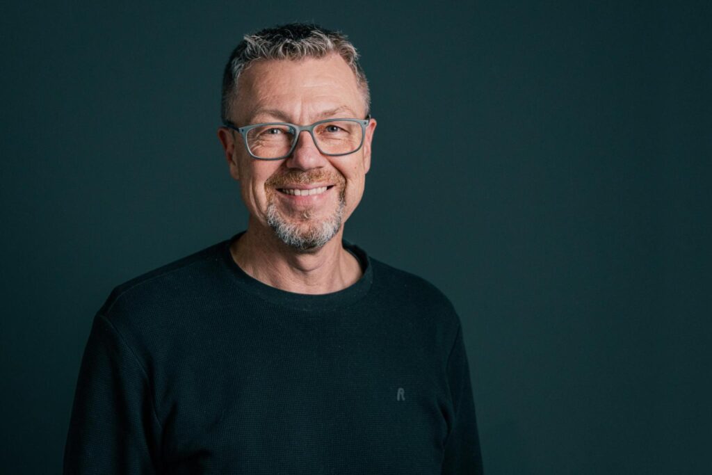 Freundlicher Mann mit Brille und grauem Bart, der bei ICF Balingen aktiv ist, in einem dunklen Pullover vor einem dunklen Hintergrund.