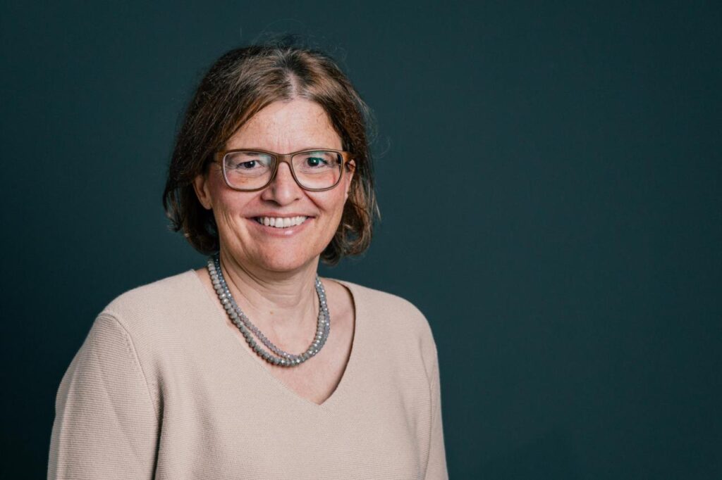 Porträt einer Frau mit Brille und Perlenkette, lächelnd, vor dunklem Hintergrund, professionell und freundlich.