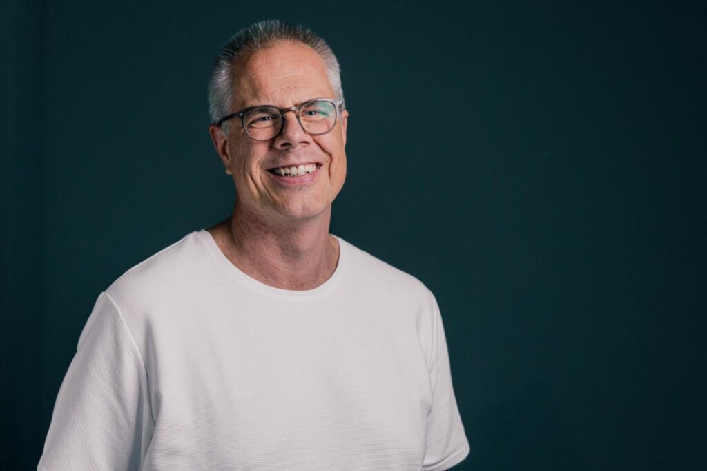 Freundlicher älterer Mann mit Brille und weißem Pullover, lächelnd vor dunklem Hintergrund, symbolisiert positive Ausstrahlung und Erfahrung.