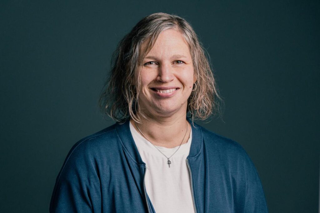 Freundliche Frau mit kurzen, grauen Haaren, lächelnd, trägt blauen Cardigan und weiße Bluse, vor dunklem Hintergrund.