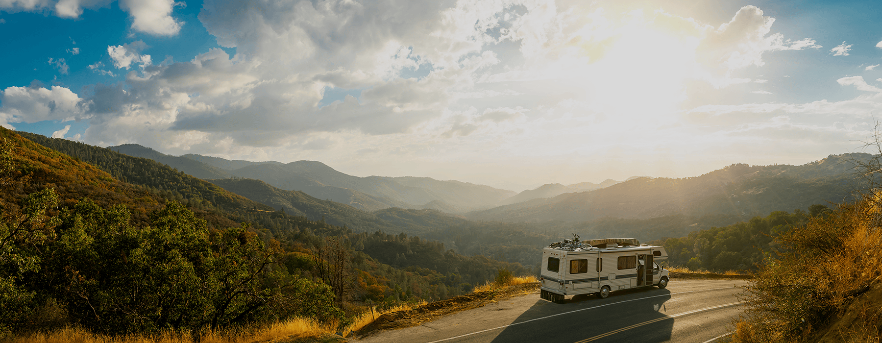 Ein Wohnmobil fährt auf einer kurvigen Straße durch eine malerische Berglandschaft bei Sonnenuntergang, umgeben von grünen Hügeln und bewölktem Himmel.