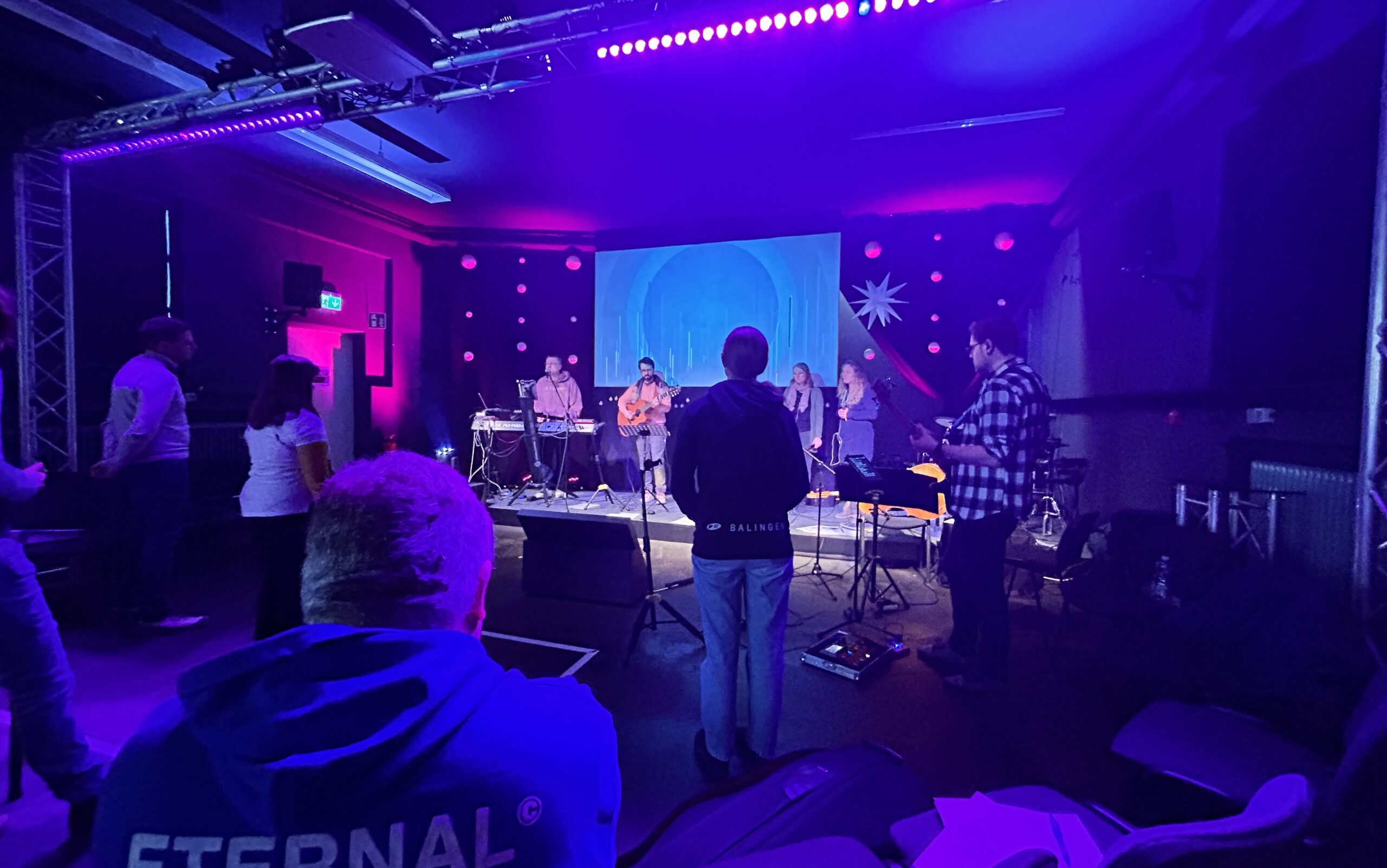 Musiker auf Bühne mit Instrumenten bei Konzert, Publikum im Vordergrund.