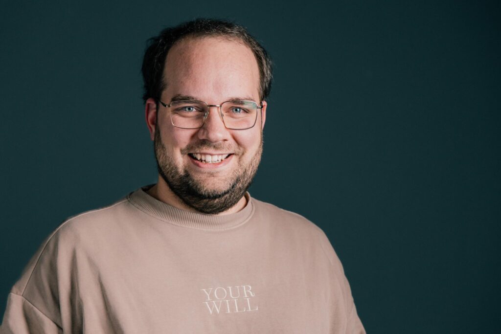 Ein Mann mit Brille und Bart, trägt ein beiges Oberteil mit dem Text "YOUR WILL".