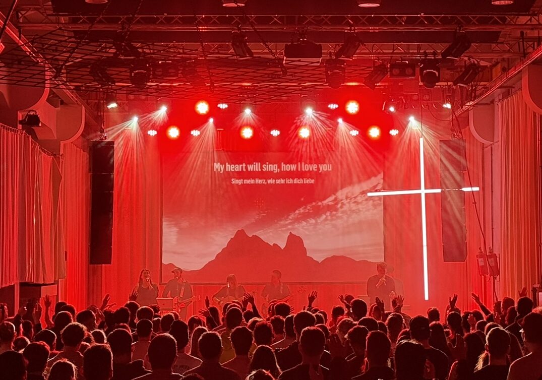 Ein Publikum sitzt in einem dunklen Raum mit roter Beleuchtung, Blick auf eine große Leinwand.