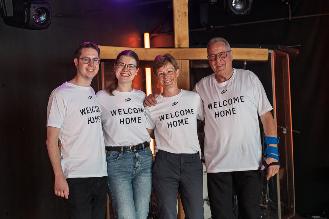 Gruppe von vier Menschen, die bei einer Veranstaltung im Rahmen von "Finde Anschluss" zusammenstehen, alle tragen T-Shirts mit "Welcome Home" und zeigen Gemeinschaft und Zugehörigkeit.