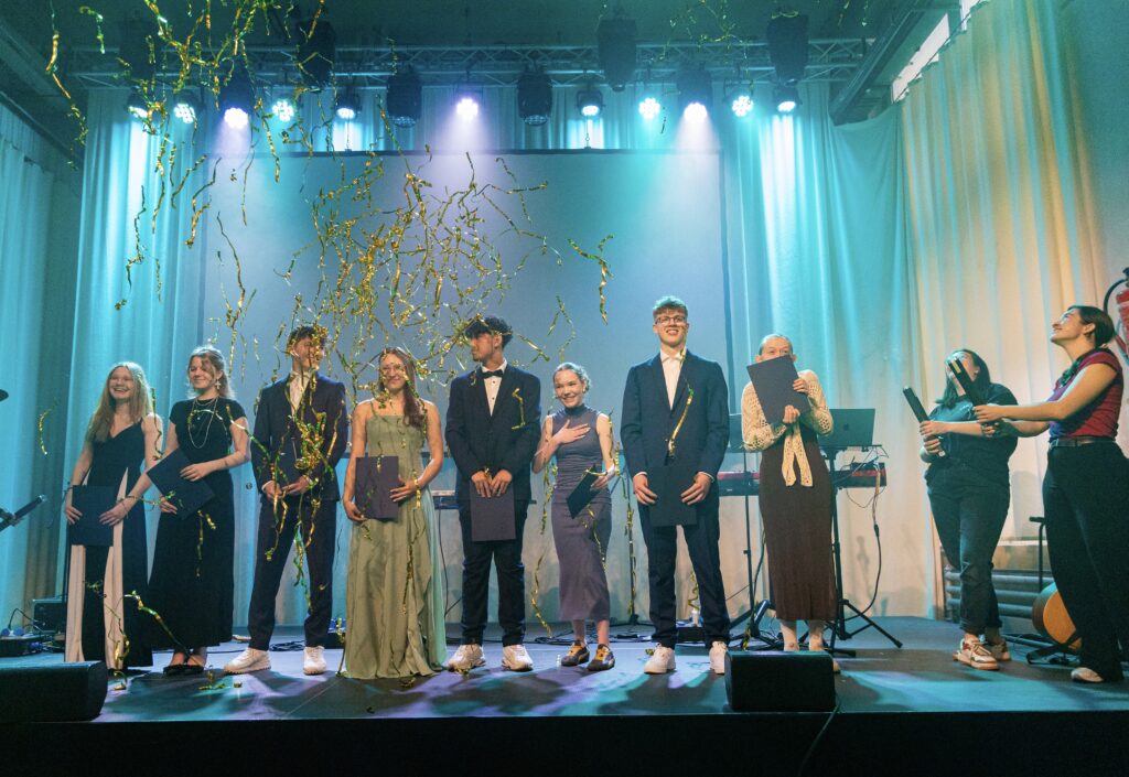 Jugendliche bei der Abschlussfeier des Jugendkollegs auf der Bühne mit Musikern und Dekorationen, festliche Atmosphäre, junge Menschen in formeller Kleidung, Feierlichkeiten, Gemeinschaftsgefühl.