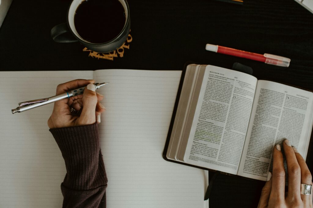 Person schreibt Notizen in ein Notizbuch neben einer offenen Bibel, einer Tasse Kaffee und einem roten Marker auf einem dunklen Tisch, symbolisiert Gebet und Studium.
