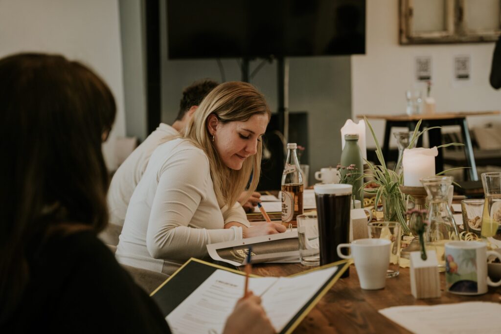 Frauen bei einem Treffen in einem modernen Raum, diskutieren und nehmen Notizen, umgeben von Pflanzen, Getränken und Notizbüchern, in einem inspirierenden kirchlichen Umfeld.