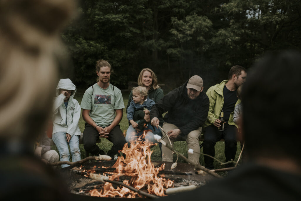 Gruppe von Menschen beim gemeinsamen Lagerfeuer im Freien, lachend und in Gespräche vertieft, umgeben von Natur, ideal für Gemeinschafts- und Outdoor-Aktivitäten.