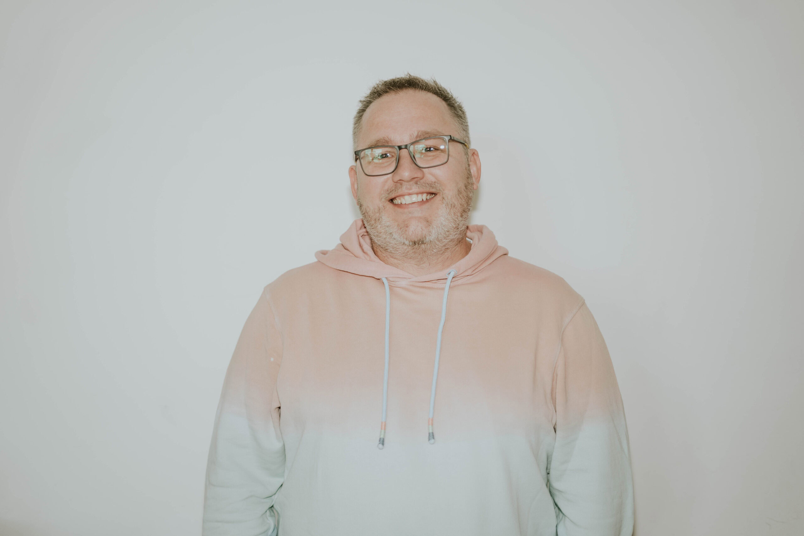 Mann mit Brille und Hoodie lächelt vor weißem Hintergrund.
