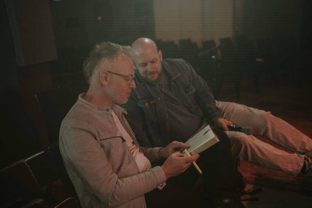 Zwei Männer sitzen in einem dunklen Raum, lesen gemeinsam in einem Buch, symbolisieren Gemeinschaft und Glauben in der Kirche ICF Berlin.