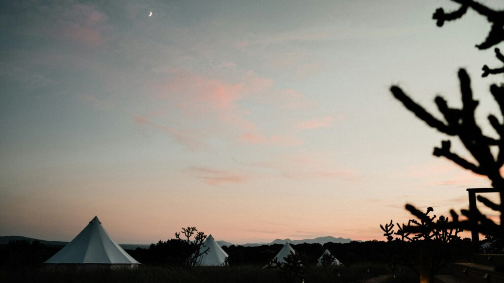 Ein Bild zeigt Zelte auf einem offenen Feld bei Sonnenuntergang, mit Bäumen im Vordergrund.