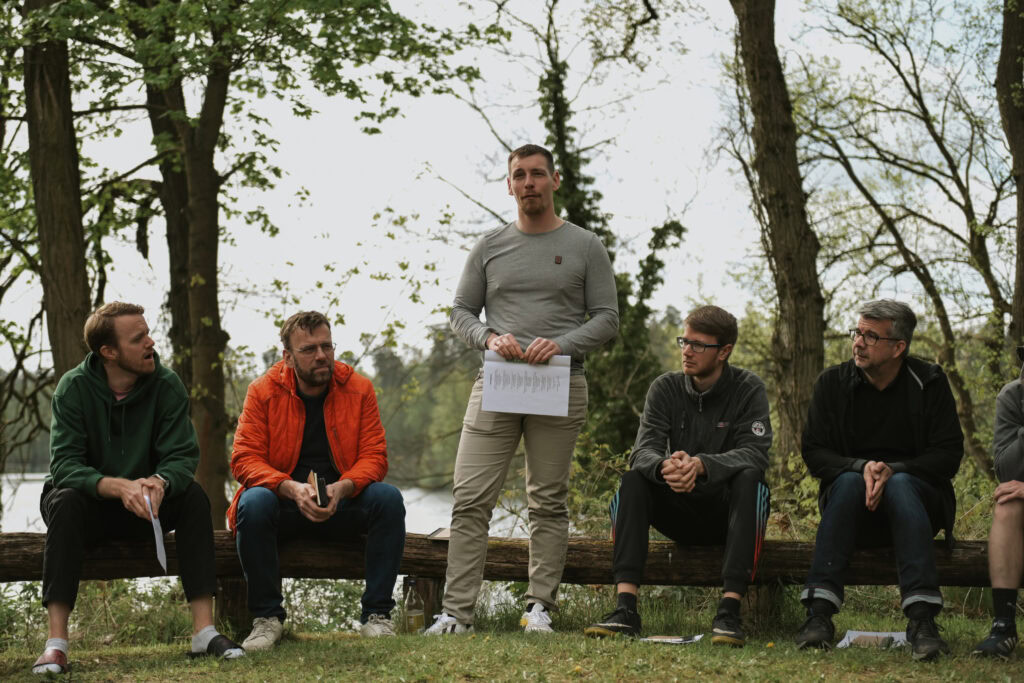 Eine Gruppe von fünf Männern sitzt im Park, einer steht und hält ein Blatt Papier.