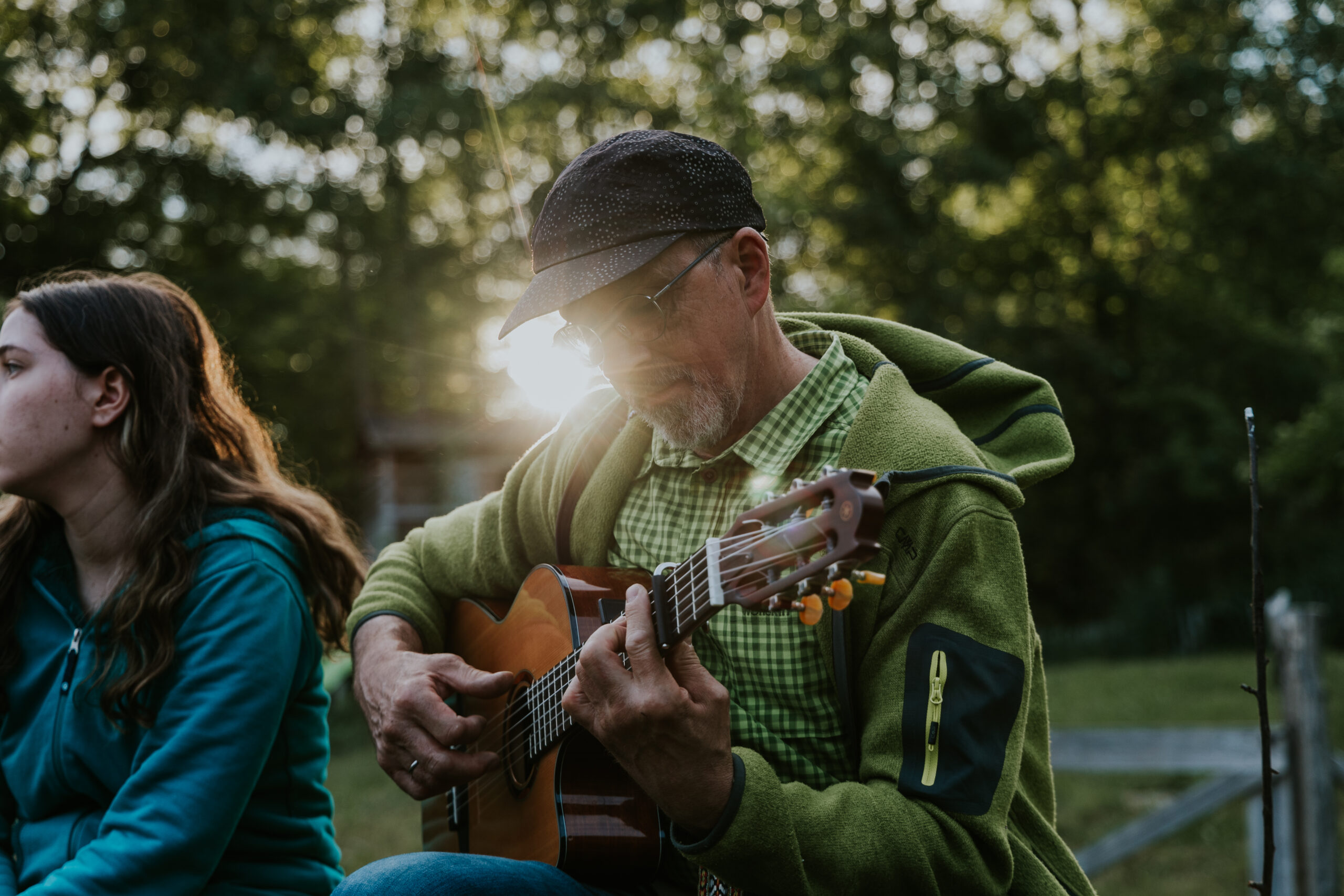Ein Mann spielt Gitarre, während er im Freien sitzt, im Hintergrund Bäume und Sonnenlicht.