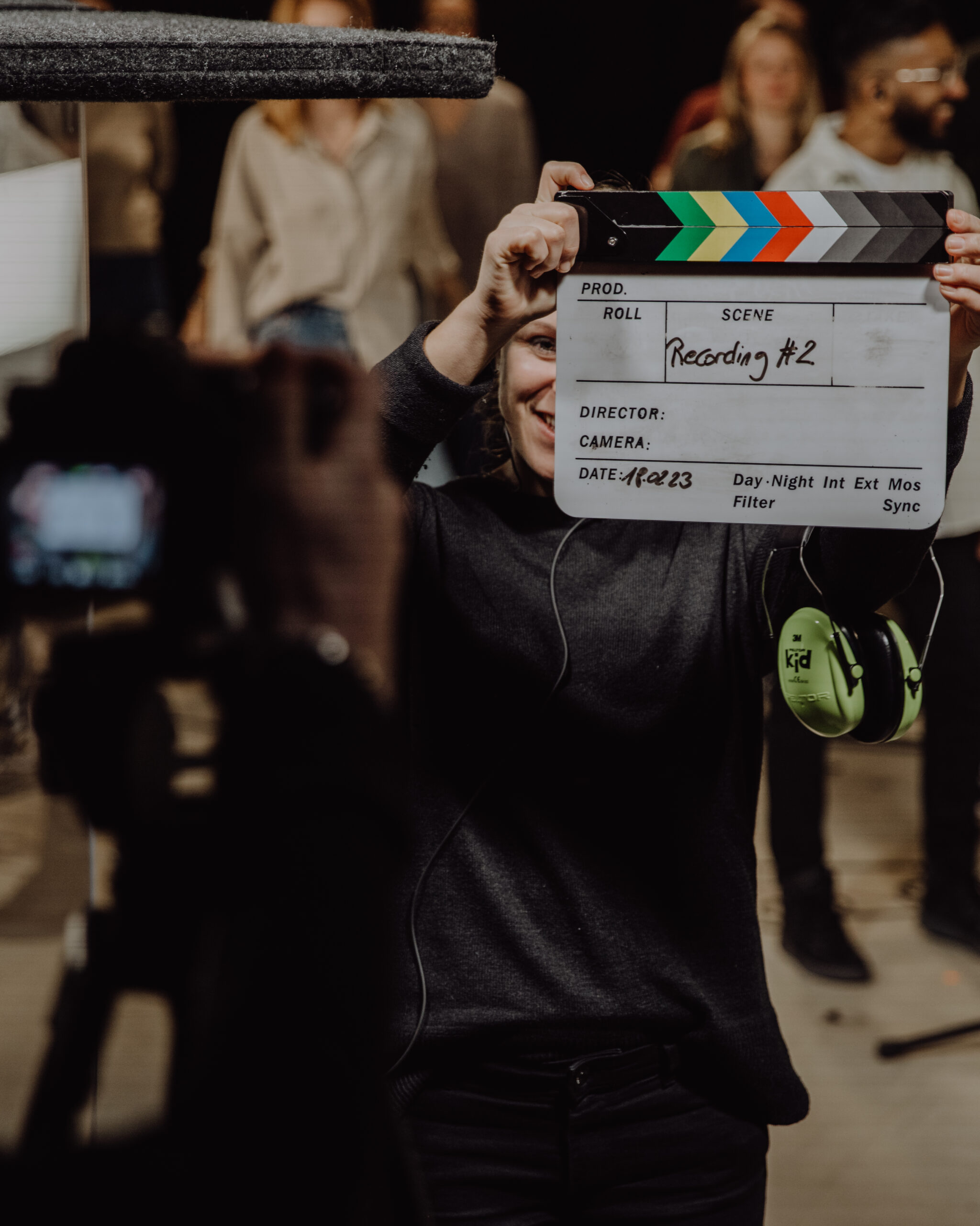 A person is holding a clapperboard labeled “Recording #2” in a group of people.