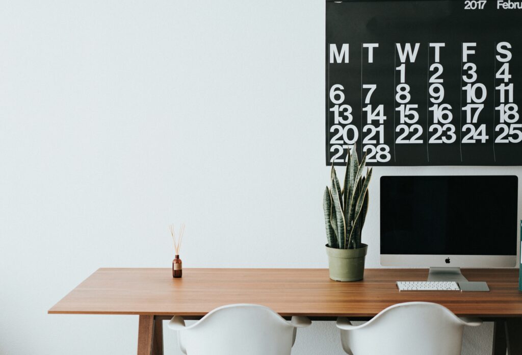 Schreibtisch mit Computer, Pflanze und Kalender an der Wand.