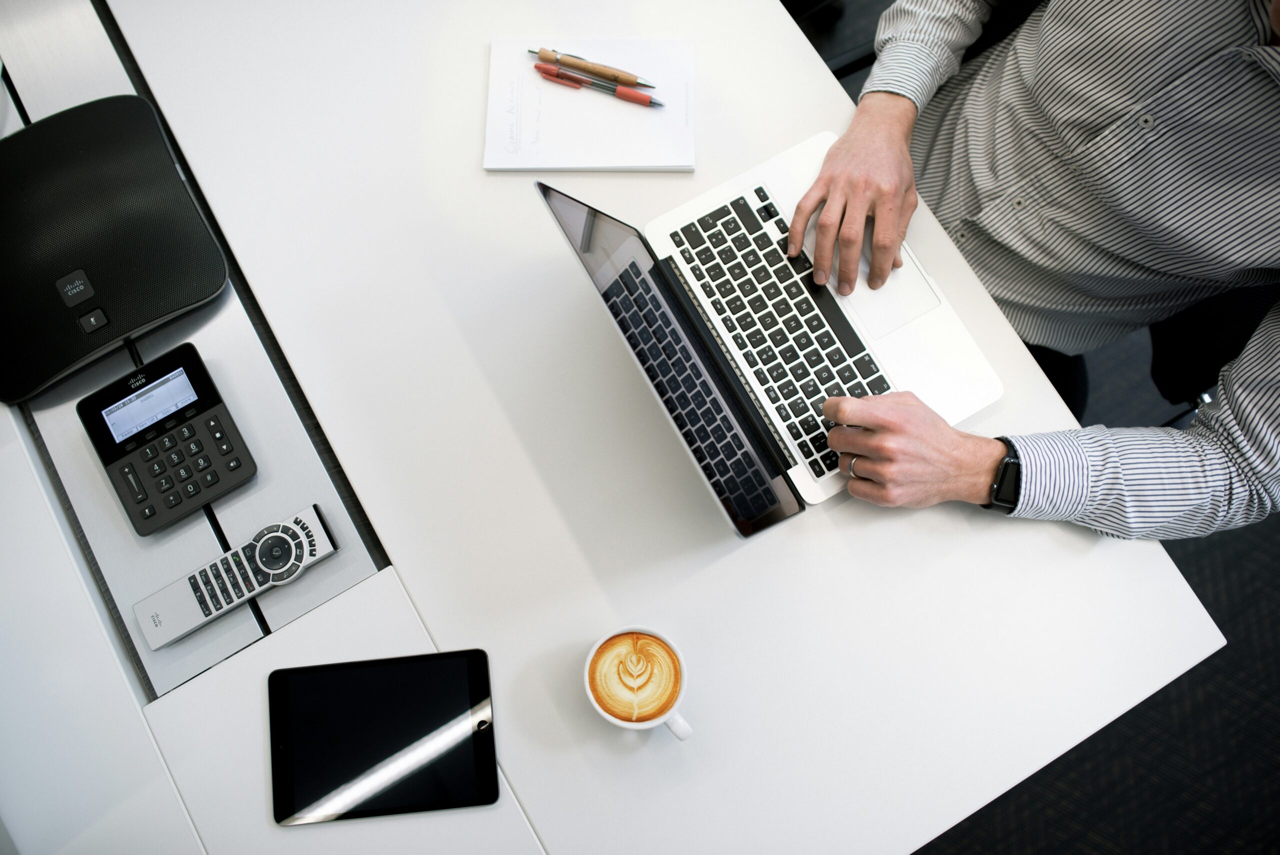 A person is working at a white desk with a laptop, smartphone, printer, notebook, and coffee.