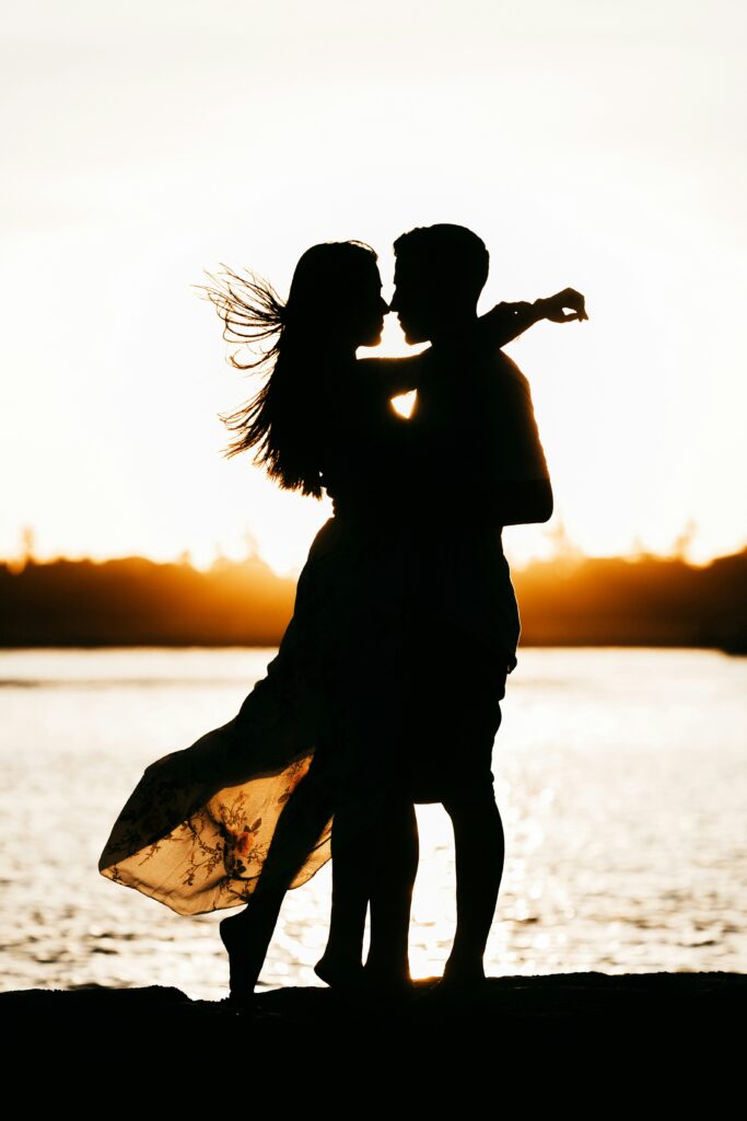 Silhouette eines verliebten Paares beim Küssen am Ufer während des Sonnenuntergangs. Romantische Stimmung, kreative Lichtsetzung, perfekte Erinnerung an besondere Momente.