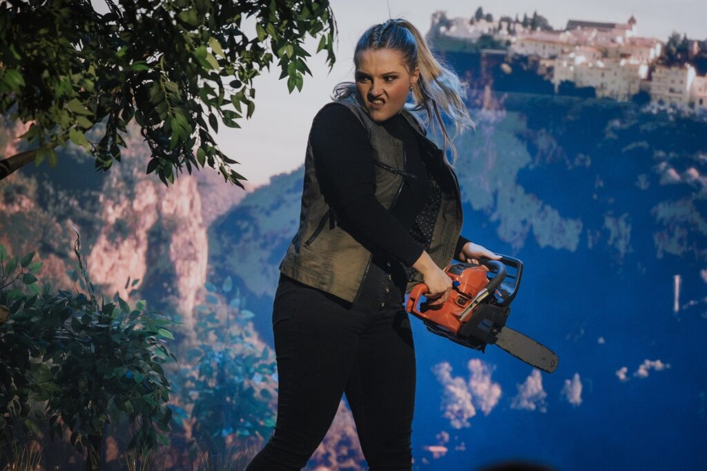 Mädchen mit dunkler Jacke und schwarzen Hosen hält eine orange Kettensäge im Freien.