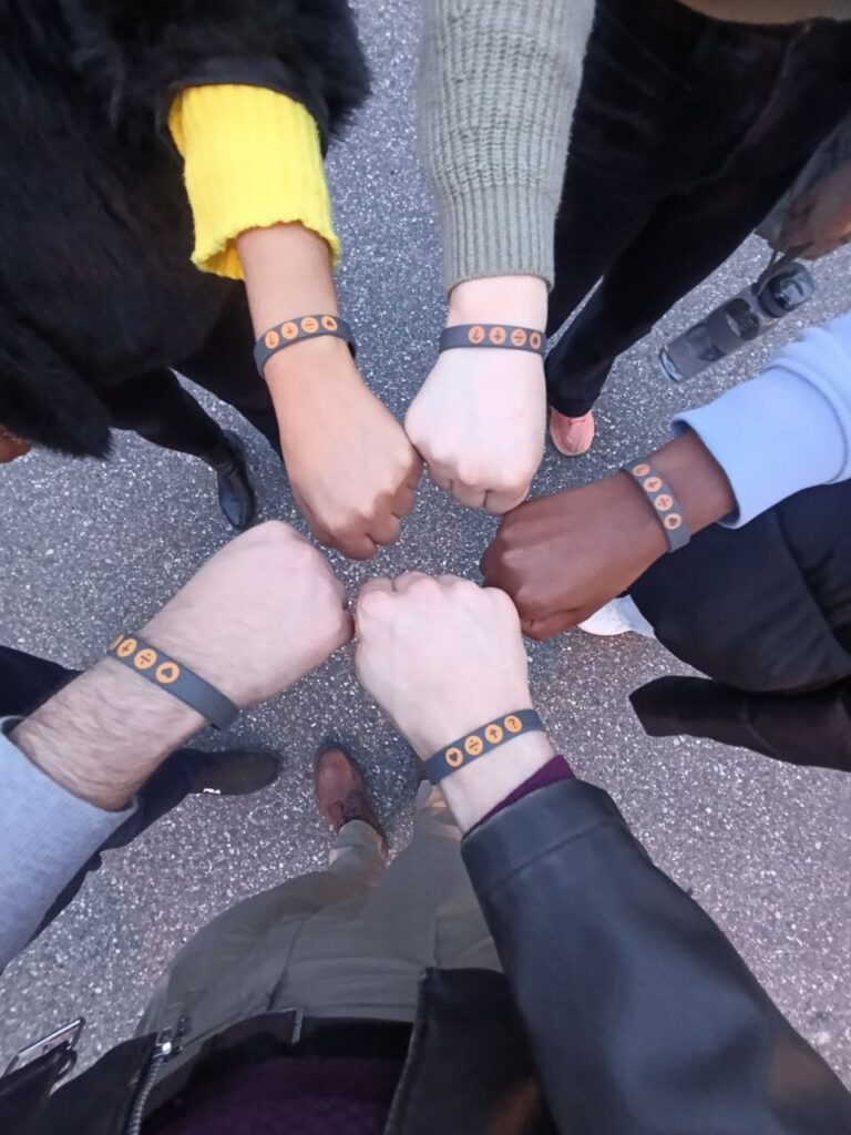 Sechs Personen zeigen ihre Handgelenke mit grauen Armbändern, die mit orangefarbenen Kreisen verz.
