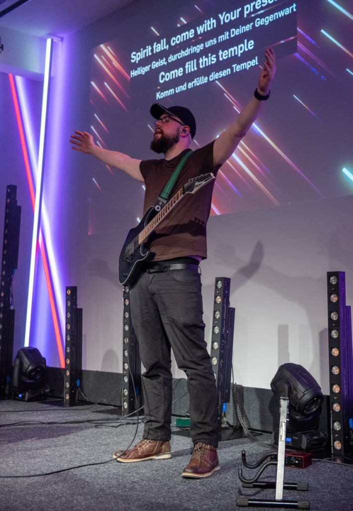 Mann mit Brille und Bart, der auf einer Bühne mit ausgestreckten Armen steht, eine Gitarre trägt.