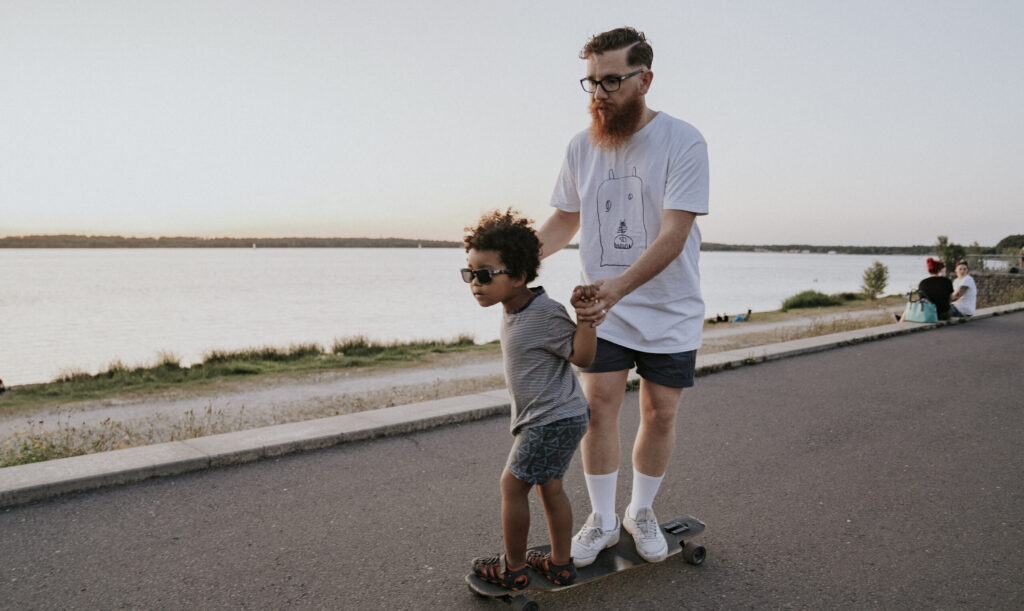 Ein Mann mit Brille, Bart und weißen T-Shirt fährt auf einem Skateboard, während ein Kind mit dunkle.