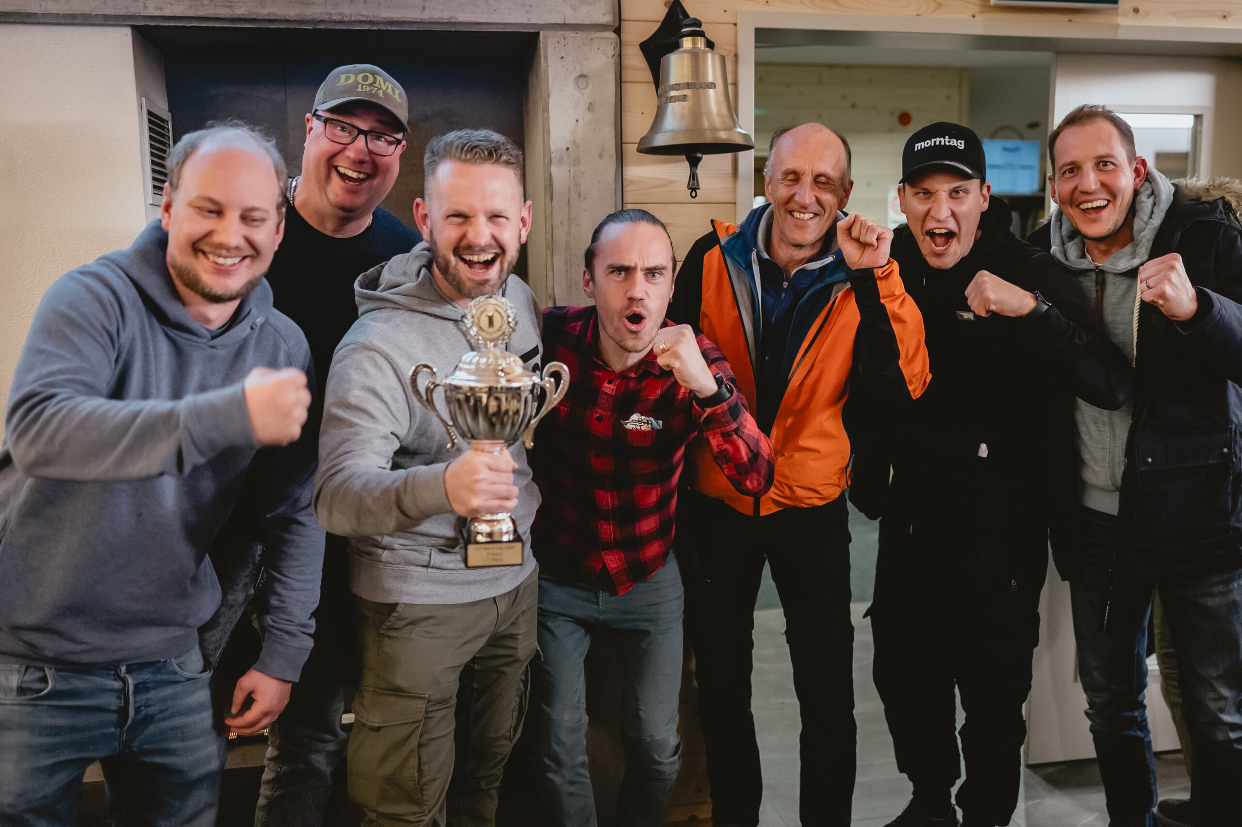 Sechs Männer stehen zusammen, einer hält einen Pokal, alle lachen und feiern.