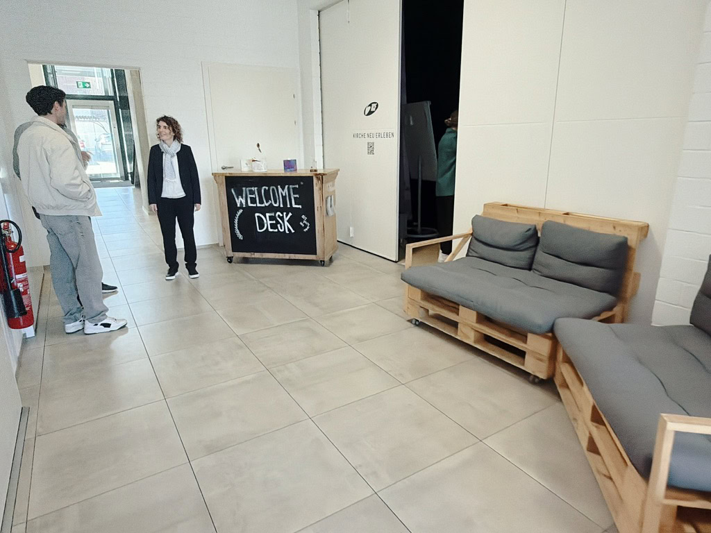 Drei Personen stehen im Eingangsbereich, zwei sprechen, eine Person steht an einem Empfangstresen.