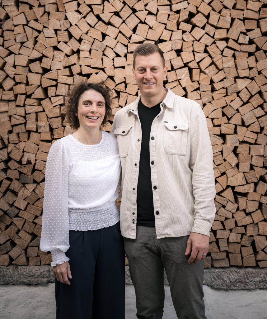 Zwei Menschen stehen vor einem Holzstapel, lächeln und schauen in die Kamera.