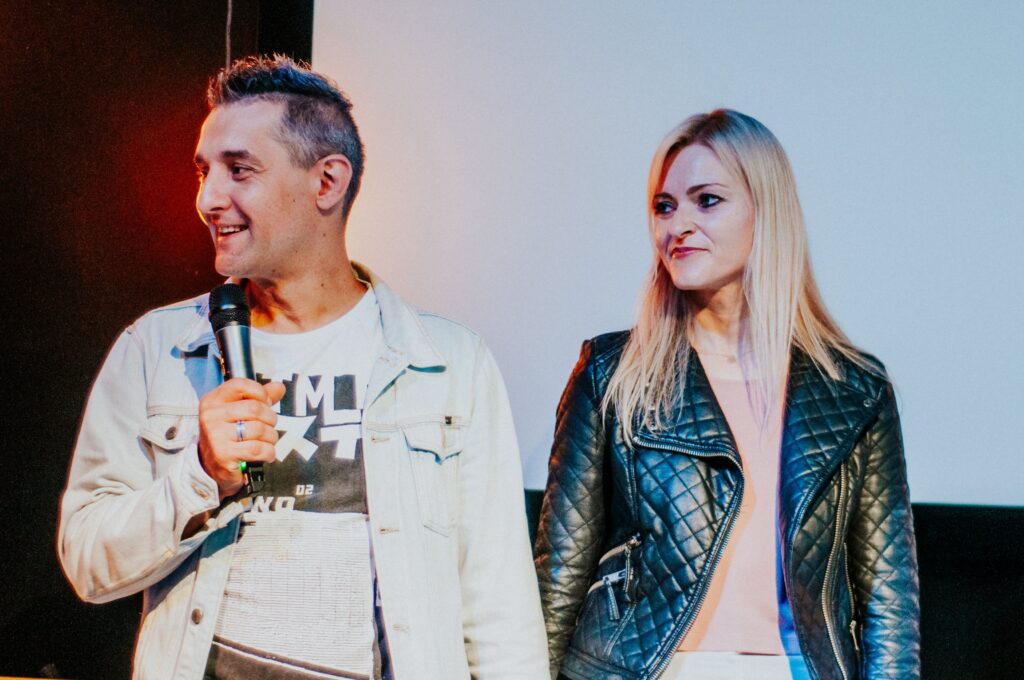 Two people standing on stage, one holding a microphone, the other looking at the first person.
