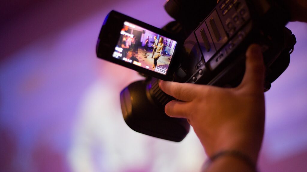 A person holds a video camera, capturing a scene in a dimly lit room with purple lighting.