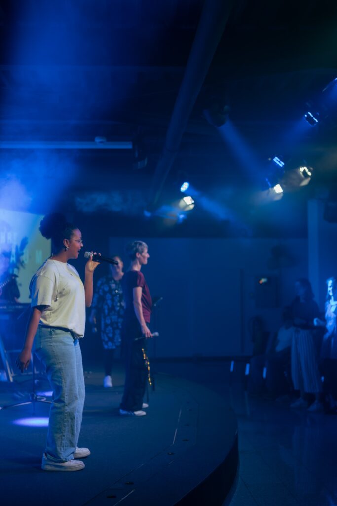 Jugendliche beim Gottesdienst mit Musik und Gebet, auf der Bühne im dunklen Raum mit blauen Lichtern, in einer modernen Kirche in Deutschland.
