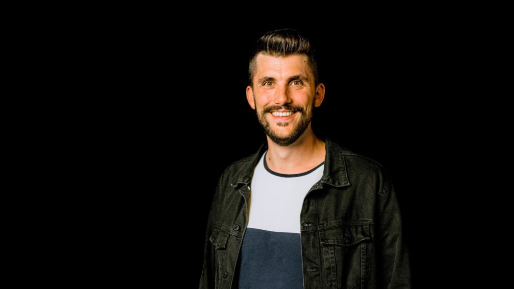 Freundlicher Mann mit dunklem Jacke und T-Shirt, der in einem hellen Raum vor schwarzem Hintergrund lächelt, symbolisiert Gemeinschaft und Glauben bei ICF Kirche.