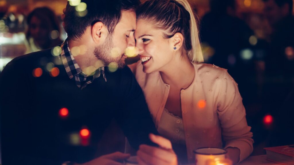 Ein verliebtes Paar bei einem romantischen Treffen, das sich in einer gemütlichen, beleuchteten Umgebung gegenüber sitzt und sich liebevoll anschaut.