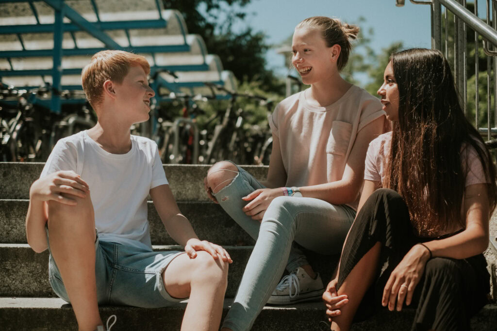 Drei Jugendliche sitzen auf Schulstufen im Gespräch, lachend und lächelnd.