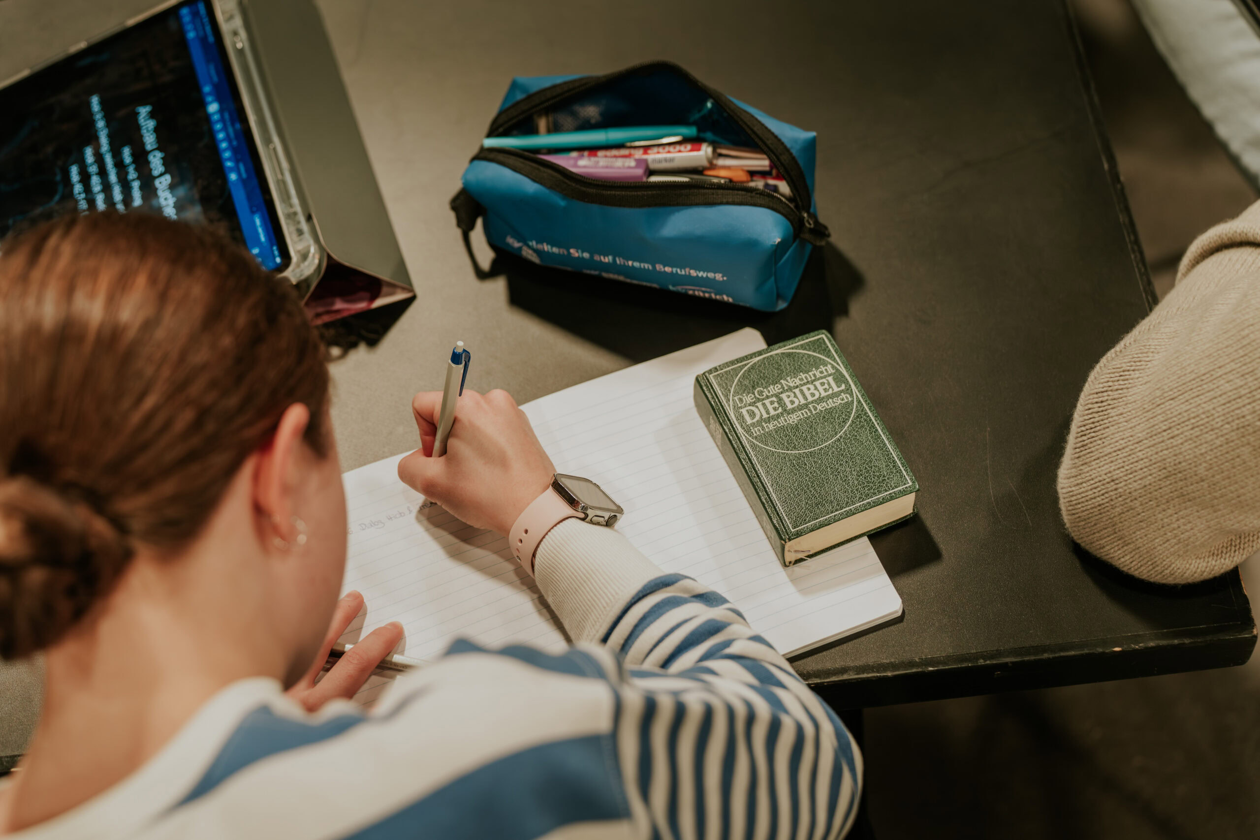 Schüler schreibt in ein Heft, neben einem Bibelbuch und einem Stift.