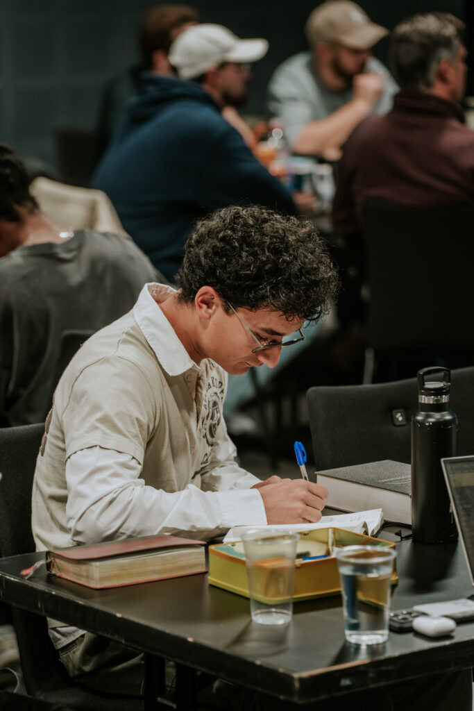 Student mit Brille schreibt in ein Heft auf einem Tisch, umgeben von Büchern und Getränken.