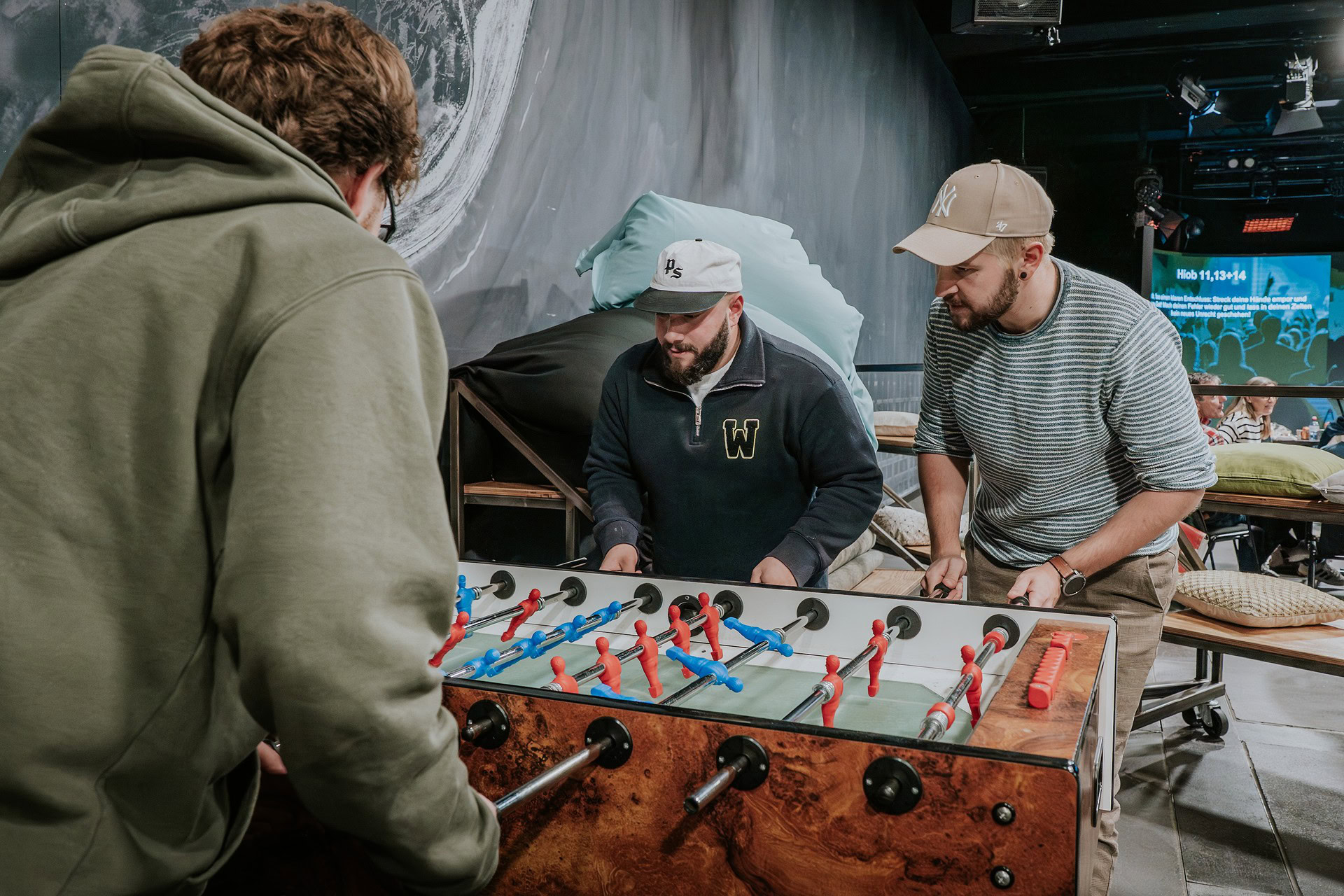 Drei Männer spielen Tischfußball in einem Raum mit dunklem Hintergrund.