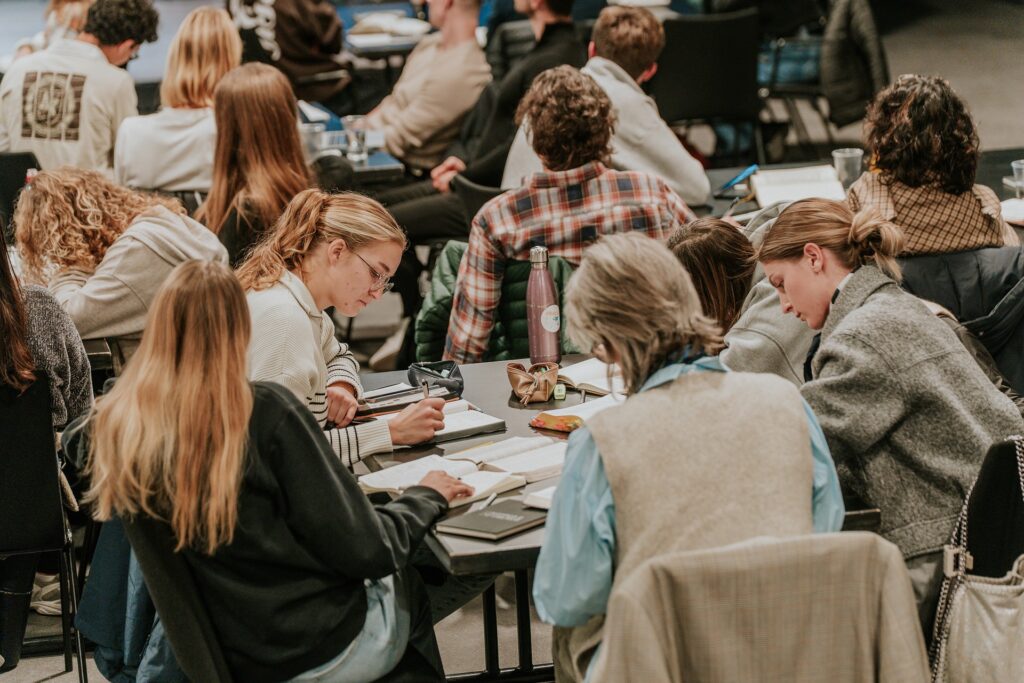 Menschen sitzen an Tischen in einem Konferenzraum, einige mit Notizbüchern und Laptops.