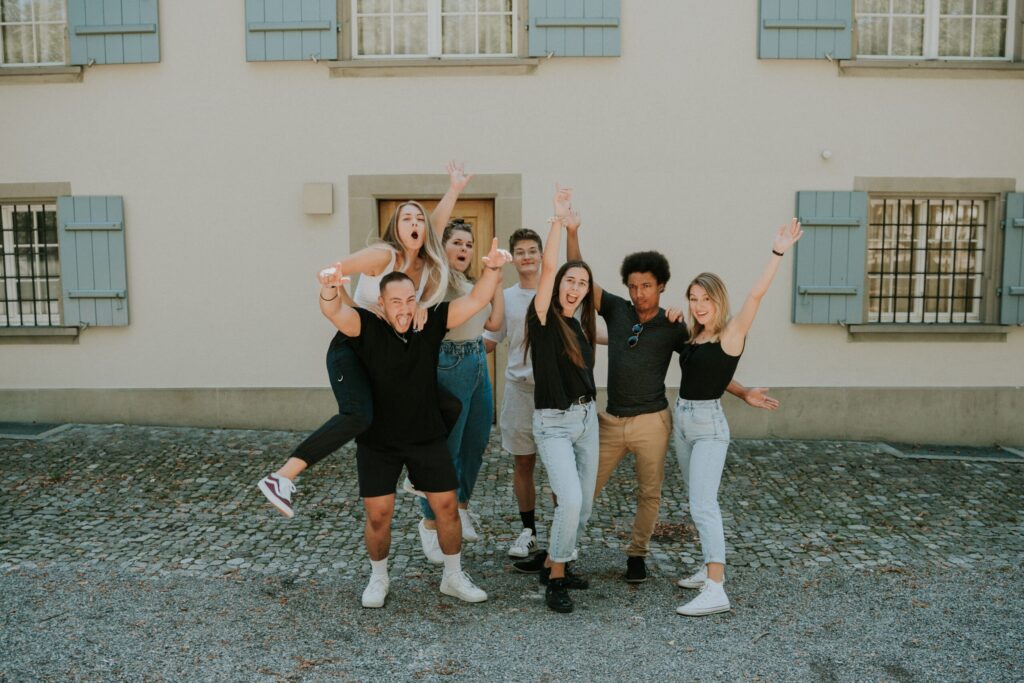 Sieben junge Menschen stehen vor einem Haus, einige heben die Arme, lachen und posieren.