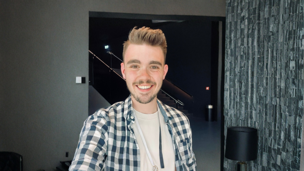 Lächelnder junger Mann mit kurzen braunen Haaren und Bart, trägt ein kariertes Hemd und ein Headset.