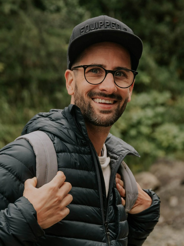 Mann trägt Brille, schwarze Mütze und Rucksack, steht im Wald.