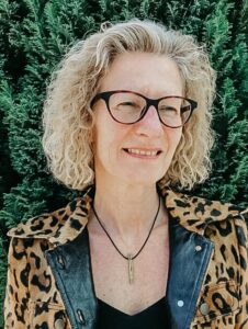 Frau mit Brille und lockigem Haar vor grünen Pflanzen.