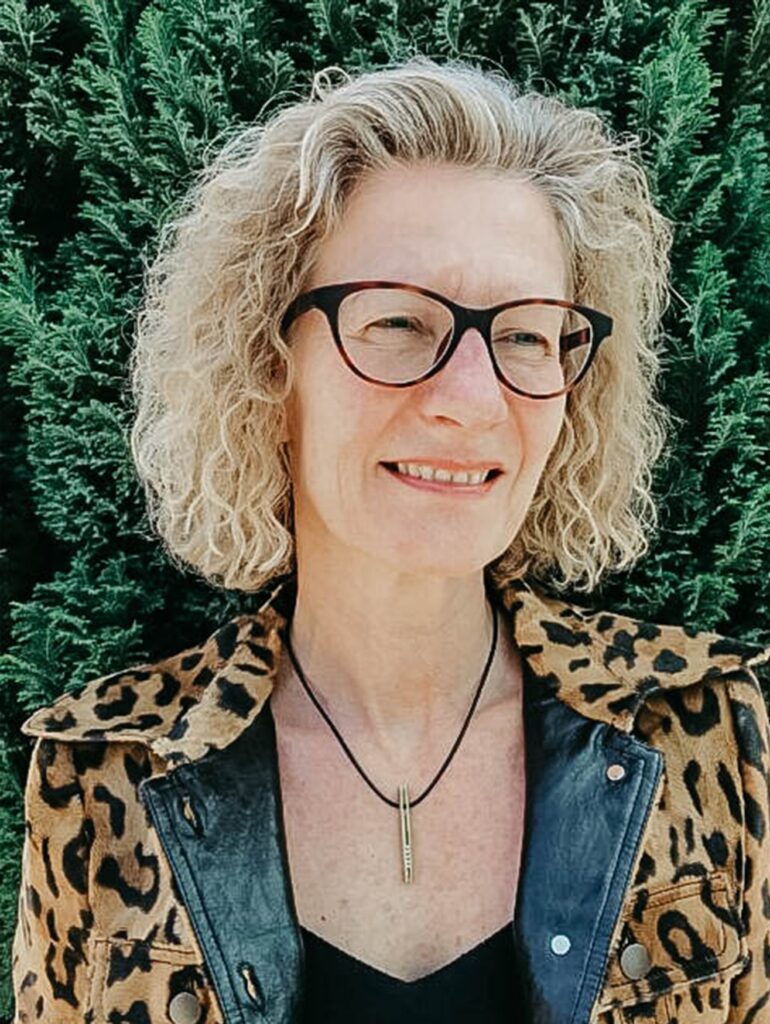 Frau mit Brille und lockigem Haar vor grünen Pflanzen.